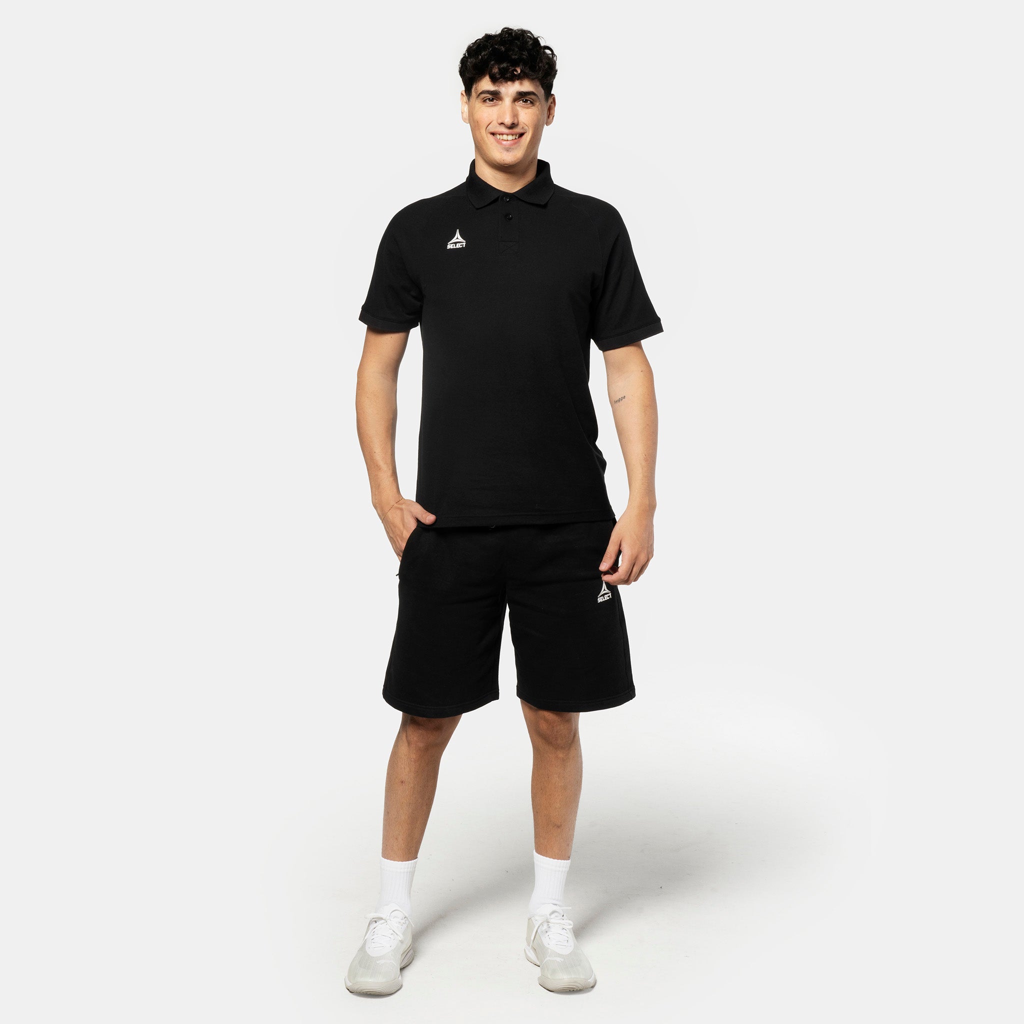 Man football player in black Sweat shorts Oxford v25 leisure wear, front view with matching polo and white sneakers.