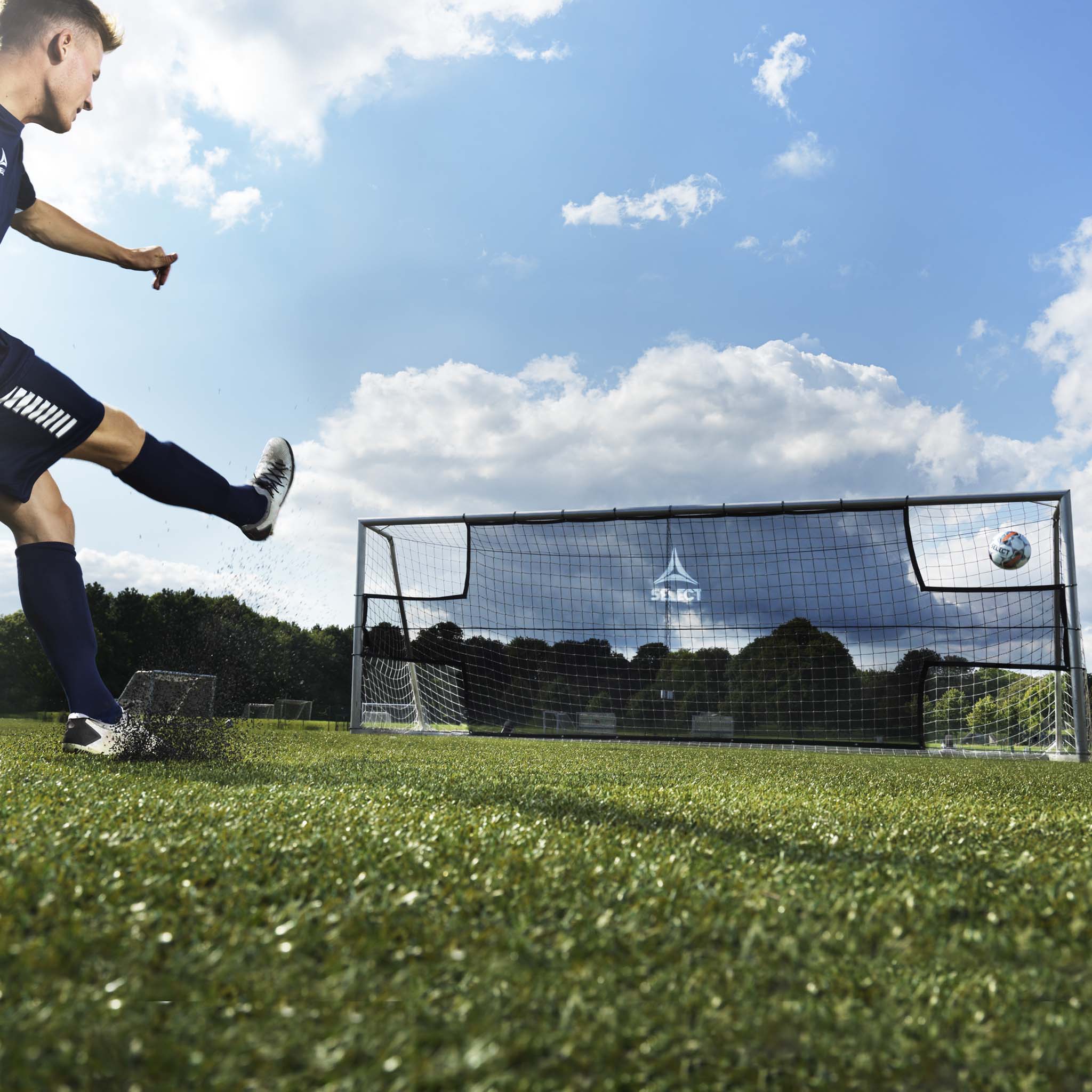 Black Goal net training equipment on grass field, front page view with football being kicked toward it under blue sky.